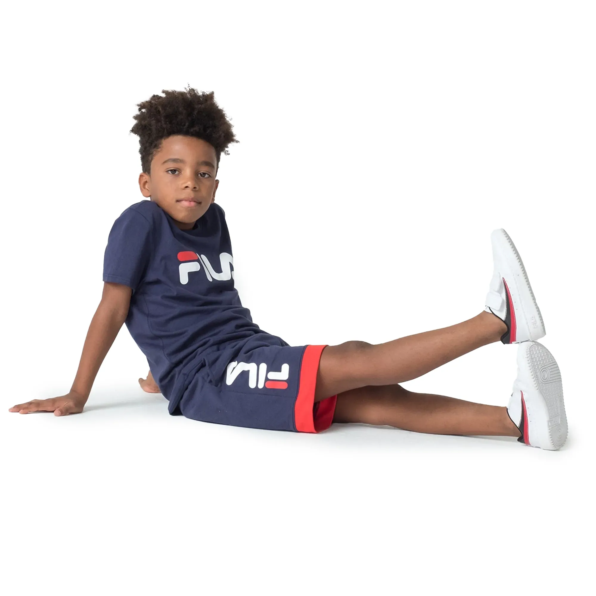 Menino sentado usando conjunto Fila juvenil camiseta e shorts marinho